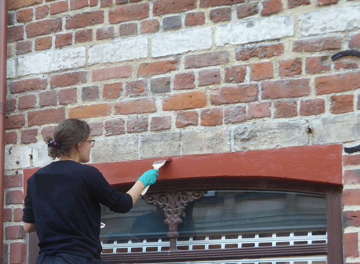 Een schilder op een stelling verft de latei boven een winkelraam in een bakstenen gevel